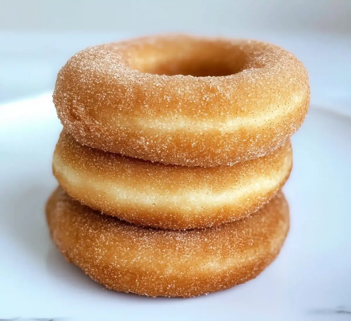 Three cinnamon-sugar coated donuts stacked on a white marble surface