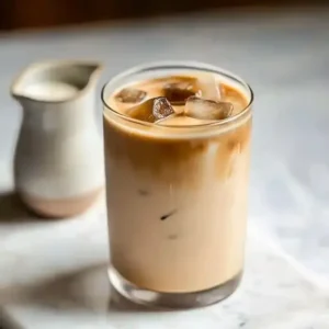 Iced brown sugar latte with ice cubes on marble surface beside cream pitcher.