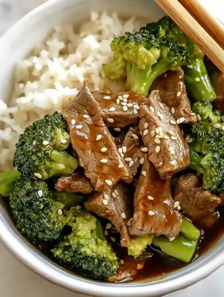 Beef and broccoli bowl with sesame seeds and chopsticks on marble surface.