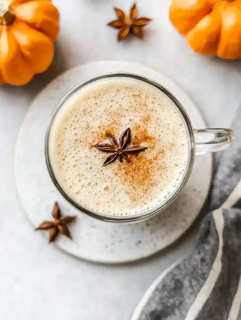 Pumpkin chai latte in a glass mug, surrounded by orange pumpkins on marble.