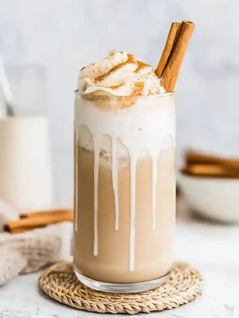 Beige beverage with whipped cream and cinnamon stick on marble surface.