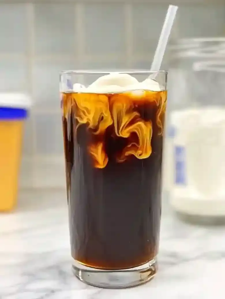 Tall glass of cold brew coffee with cream swirled on marble surface.