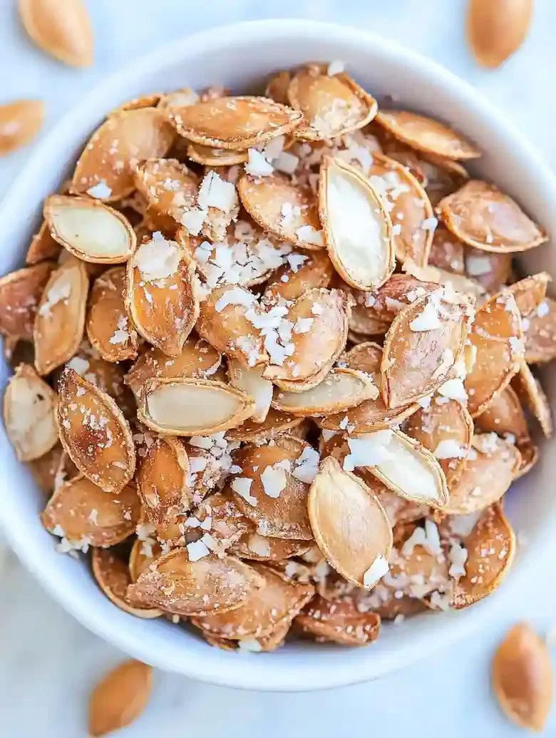 Bowl of roasted pumpkin seeds coated in garlic and cheese on marble surface.
