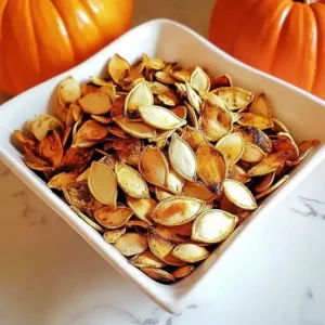 White dish with golden-brown roasted pumpkin seeds on a marble surface.