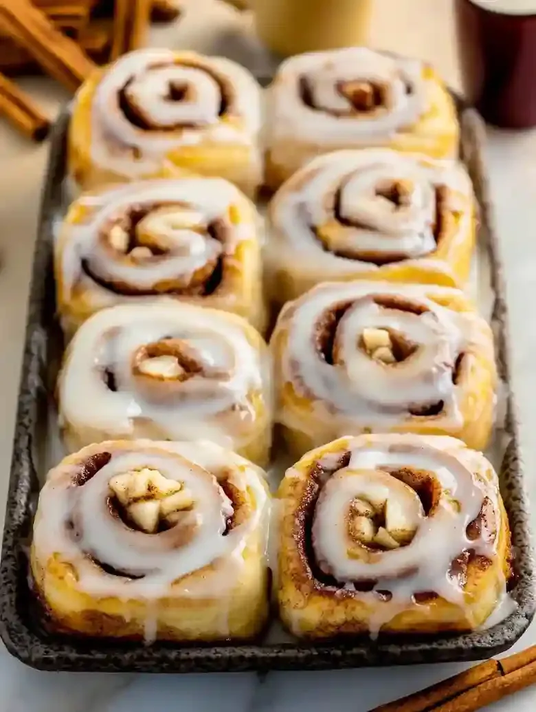 Golden-brown cinnamon rolls with icing, apples, and cinnamon on marble.